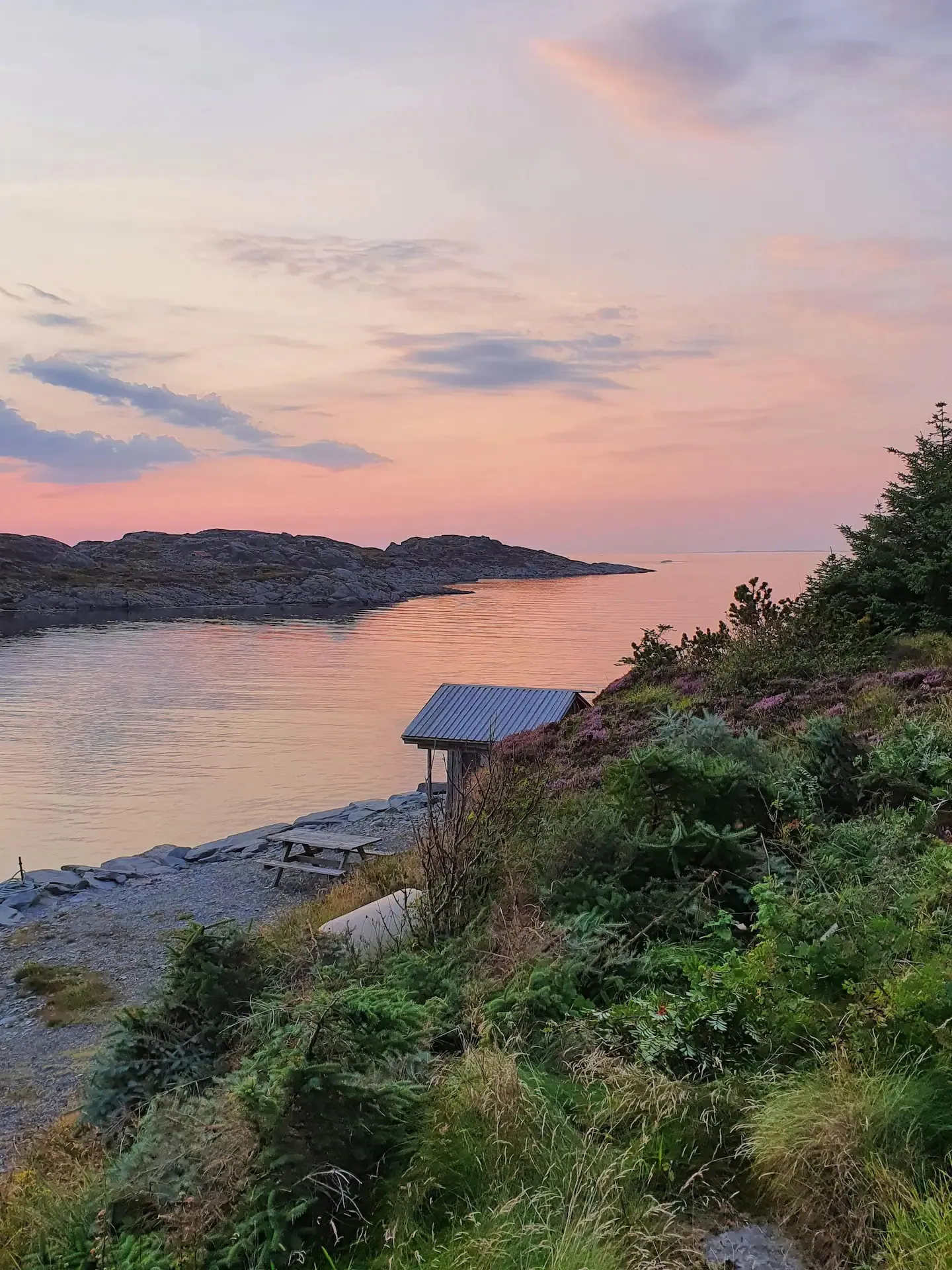 Solnedgang i rosa og oransje over viken nedenfor huset, med naust og benker ved vannkanten